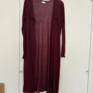 Vinröd lång kofta från Vila - Lång och tunn vinröd kofta från Vila med öppen front och långa ärmar. Perfekt för lager-på-lager och ger en chill vibe till din outfit. Enkel design utan knappar eller dragkedja, i ett lätt och mjukt material.