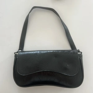 Helt ny svart axelväska i skinnimitation - Stilren svart axelväska i skinnimitation med snyggt krackelerad yta. Väskan har en böjd form, magnetlås och ett huvudfack med dragkedja inuti. Helt ny.