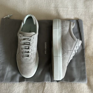 Grå mockasneakers från Axel Arigato - Snygga sneakers i ljusgrå mocka från Axel Arigato med klassisk siluett och platt vit sula. De är endast använda en gång och säljs då dom tyvärr är för små. Både original box och skopåse ingå. Kartongen är det sista originalet av den gamla kartongen som finns. De köptes för 2650 men säljs för 2000, priset går att diskutera💞