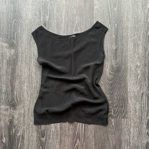 Svart ärmlös silkes linnetopp  - En stilren svart linnetopp med bred båturringning och ärmlös design. Toppen har en slät yta och tre små knappar i sidan nertill som snygg detalj. Perfekt att matcha med jeans eller kjol för en clean look.