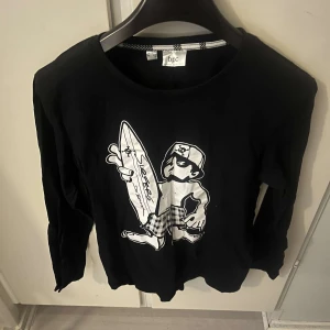 Svart långärmad tröja med surf-tryck - Cool svart långärmad tröja från bpc med ett stort vitt tryck av en tecknad surfare på framsidan. Tröjan har rund halsringning och är gjord i mjuk bomull, perfekt för en avslappnad streetstil.