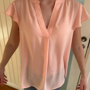 Ljusrosa blus från Calvin Klein - Superfin ljusrosa blus från Calvin Klein med v-ringning och korta ärmar. Blusen har en lös passform och är tillverkad i ett lätt, flowy material som känns skönt