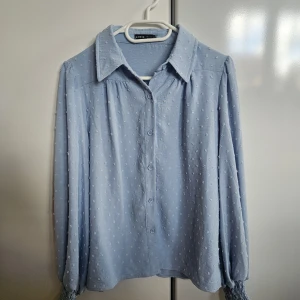 Ljusblå skjorta med struktur från SHEIN - Supersöt ljusblå skjorta från SHEIN med små broderade prickar och knappar framtill. Skjortan har långärmade puffiga ärmar med smockade muddar och klassisk krage. Perfekt för dig som gillar en feminin och trendig look.