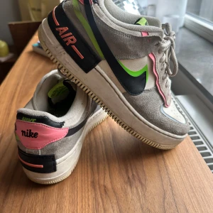 Nike Air Force 1 sneakers i grå och neon - Nike Air Force 1 sneakers med grå mocka, vita partier och detaljer i neonrosa, limegrönt och svart. Swoosh-logga i svart med grön kant, rosa detaljer vid hälen och AIR-tryck på sulan. Perforerad tå, snörning och klassisk platt sula. Perfekt för dig som gillar färg och streetstyle.