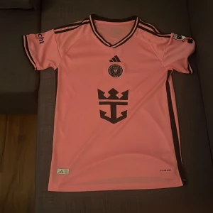 Rosa Inter Miami fotbollströja Adidas - Snygg rosa Inter Miami fotbollströja från Adidas med svarta detaljer och klubbmärke på bröstet. Kortärmad modell i lätt och ventilerande material, perfekt för träning eller match. Klassiska Adidas-ränder på axlarna och svart V-ringad krage.