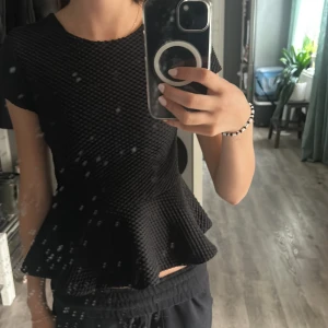 Svart peplumtop från H&M - Säljer en svart peplumtop från H&M med korta ärmar och coolt strukturerat tyg. Toppen har en markerad midja och volangig nederdel som ger en snygg siluett. Perfekt för dig som gillar unika detaljer och vill sticka ut lite extra.