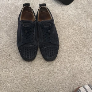 Svarta Louboutin sneakers med nitar - Säljer ett par svarta sneakers från Christian Louboutin med coola svarta nitar på tån och klassisk röd sula. Skorna har snörning och är gjorda i skinn med en rund tå. Perfekt för dig som vill sticka ut med en edgy vibe.