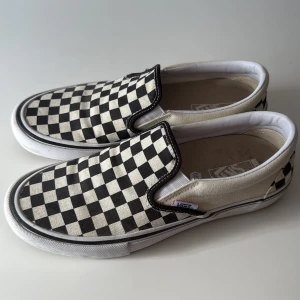 Vans slip-on checkerboard sneakers - Klassiska Vans slip-on sneakers med svart och vit rutigt checkerboard-mönster. Skorna har en låg profil, platt sula och är tillverkade i bomullscanvas. Storlek 42 (US 9). Innermått 27cm. Använda vid två tillfällen. 