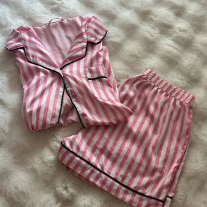 Rosa och vita randiga pyjamasshorts och tröja - Säljer ett par pyjamasshorts och tröja med rosa och vita ränder, elastisk midja och svarta kantdetaljer. De är loose fit och har en mjuk känsla, perfekta för chill hemma. Materialet är bomull och mönstret är klassiskt randigt.