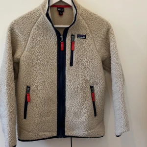 Beige fleecejacka från Patagonia - Säljer en beige fleecejacka från Patagonia med marinblå detaljer och röda dragkedjor. Jackan har tre fickor med dragkedja, hög krage. Perfekt för kalla dagar och chill stunder utomhus.