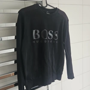 Svart slim fit tröja från Hugo Boss - Svart långärmad tröja från Hugo Boss med stor BOSS-logga framtill. Tröjan har en stilren design med ribbad krage och muddar, samt en diskret panel i mörkare ton över axlarna. Perfekt för dig som gillar klassisk streetwear med premiumkänsla. En jävligt snygg tröja som tyvärr är för liten för mig. Storlek small