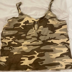 Beige camouflage linne med cut-out - Säljer ett linne i beige och bruna camouflagefärger med smala axelband och en liten cut-out detalj framtill. Toppen har en loose passform och är tillverkad i mjuk polyester som känns skön mot huden. Perfekt för dig som gillar streetstyle och vill sticka ut.