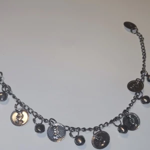 Silverfärgat armband med mynt - Snyggt armband i silverfärg med kedja och hängande myntberlocker samt små klot. Mynten har präglade motiv som ger en cool och unik look. Perfekt för dig som gillar detaljer och vill sticka ut lite extra.