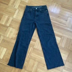 Svarta wide jeans från Never Denim - Säljer ett par svarta wide leg jeans från Never Denim, modell JP Wide Black. Byxorna har hög midja, raka ben med rå kant nedtill och klassisk femficksdesign. Materialet är 100% bomull för en skön och lite styvare känsla. Perfekt för dig som gillar en avslappnad och trendig look.