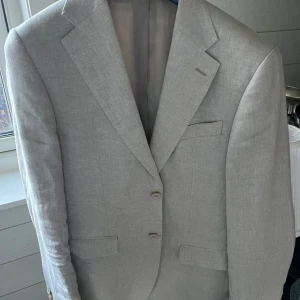 Beige kavaj från Bläck i linne - Snygg ljusgrå kavaj från Bläck med klassisk passform och två knappar framtill. Tillverkad i linne, perfekt för dig som gillar stilren och skandinavisk design. Fina detaljer som knappslå vid ärmslut och diskreta fickor fram. Passar dig som vill ha en fräsch och modern look.