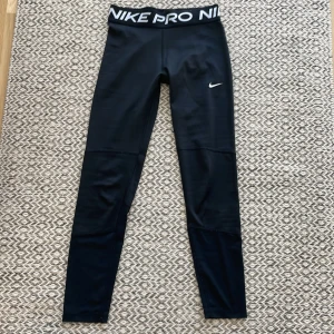 Nike Pro svarta träningstights barn 146-156 - Nike Pro leggings i svart med bred resår i midjan med vit logga. Tillverkade i Dri-FIT material som andas och transporterar bort fukt. Tight passform och stretchig polyester, perfekt för gym eller sport. Snyggt minimalistiskt utseende med liten Nike Swoosh på benet.