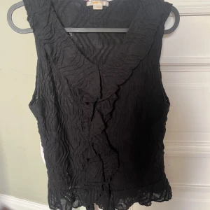 Svart volangblus från Gina Tricot - Säljer en svart blus från Gina Tricot med snygga volangdetaljer längs halsringningen och framtill. Blusen är ärmlös och har ett transparent, vågigt tyg som ger en cool struktur. 