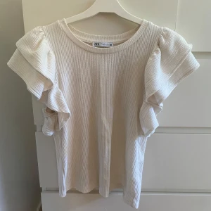 Vit ribbad topp med volangärmar från Zara - Supersöt vit topp från Zara med ribbad struktur och stora volangärmar som ger en extra touch. Rund halsringning och mjukt material som känns skönt mot huden. Perfekt för dig som gillar en mjuk och stilig tröja!