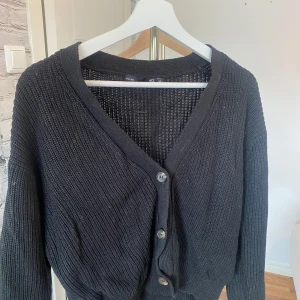 Svart stickad kofta från 157 - Svart stickad kofta från 157 med v-ringning och tre knappar framtill. Koftan har långärmade ärmar och ett grovt ribbat mönster. Perfekt att slänga över en t-shirt när det är lite kyligt ute.