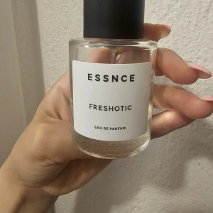 ESSNCE Freshotic Eau de Parfum - Minimalistisk parfymflaska från ESSNCE med doften Freshotic. Flaskan är cylindrisk i genomskinligt glas med svart matt kork och vit etikett med svart text. Innehållet är ljusgult och flaskan har en stilren, modern look.