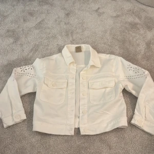 Vit jeansjacka med broderade detaljer - Snygg vit jeansjacka från Dixie med broderade blommor och hålmönster på ärmarna. Jackan har klassisk krage, två bröstfickor med lock och knappar framtill. Materialet är kraftig bomull och passformen är normal. Perfekt för dig som gillar unika detaljer.