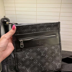 Svart axelväska - Snygg svart axelväska med klassiskt monogrammönster och silverfärgade dragkedjor. Väskan har justerbar axelrem, flera fack och är tillverkad i skinn med textilinsida. Perfekt för dig som vill ha en stilren och praktisk väska med lyxig känsla.