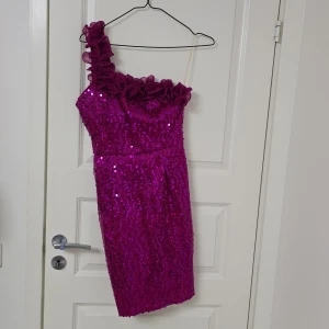Glittrig rosa one shoulder klänning - Säljer en kort, glittrig rosa klänning med paljetter och en snygg one shoulder-design. Axelbandet har volangdetaljer som ger extra wow-effekt. Perfekt för dig som vill sticka ut och glänsa på festen! Klänningen är figurnära och har en cool, modern vibe.