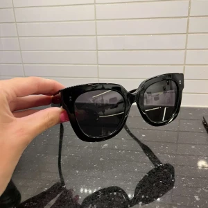 Svarta CHIMI solglasögon med fodral - Snygga svarta solglasögon från CHIMI med breda bågar och fyrkantig form. Glasen är mörka och bågarna är tillverkade i blankt syntetmaterial. Kommer med ett stilrent vitt CHIMI-fodral. Perfekt för dig som gillar statement-accessoarer.