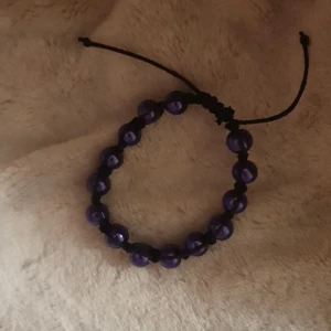 Svart armband med lila pärlor - Snyggt handgjort armband med svarta snören och glansiga lila pärlor. Justerbar knytning så det passar de flesta handleder. Perfekt accessoar för dig som vill sticka ut med lite färg och coola detaljer.