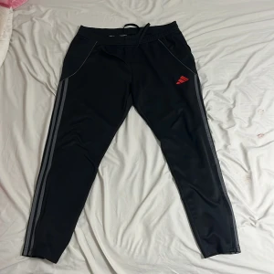 Svarta Adidas träningsbyxor XL - Svarta träningsbyxor från Adidas i slim fit med klassiska grå ränder längs benen och röd Adidas-logga på höften. Byxorna har snörning i midjan och är gjorda i mjukt syntetmaterial som är perfekt för sport och chill. Snygg, modern look med smal passform.