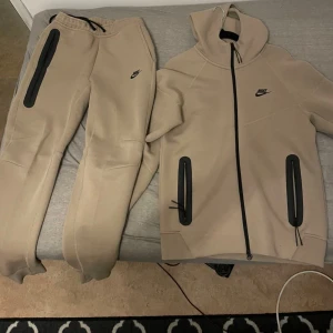 Beige Nike hoodie med svarta detaljer - Snygg beige hoodie från Nike med svarta dragkedjor och logga på bröstet. Hoodien har huva och långa ärmar, samt två praktiska fickor med svarta dragkedjor. Perfekt för chill eller träning, stilren och modern design. Vi kan diskutera om priset 