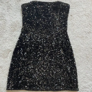 Svart paljettklänning från Lindex - Säljer en svart, axelbandslös kortklänning från Lindex i storlek S. Klänningen är täckt med glittriga paljetter som ger en riktigt festlig vibe. Den har en figurnära passform och är perfekt för dig som vill sticka ut med lite extra bling. Supersnygg till fest eller bal! Aldrig använd, prislappen kvar.
