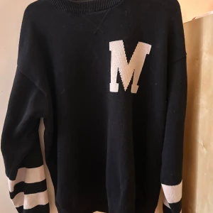 Svart stickad tröja med M-tryck - Cool svart stickad tröja med stor vit M på bröstet och vita ränder på ärmarna. Rund hals och långärmad modell, perfekt för dig som gillar sportig och avslappnad stil. Passar lika bra till jeans som till mjukisbyxor.
