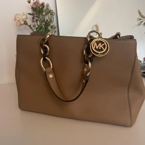 Beige handväska från Michael Kors - Snygg beige handväska från Michael Kors med guldfärgade detaljer och MK-logga. Väskan har rejäla handtag med kedjor i guld och brunt, perfekt för att bära med sig allt viktigt. Stilren design som passar till många outfits.