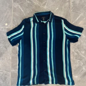 Randig blå skjorta från New Look - Snygg kortärmad skjorta från New Look i storlek S. Skjortan har ett fräscht randigt mönster i olika blå nyanser och vitt, klassisk krage och knappar framtill. Perfekt för dig som gillar en chill och stilren look.