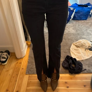 Svarta jeansbyxor med slits - Säljer ett par svarta jeansbyxor med snygga slitsar framtill vid bensluten. Byxorna har en rak passform och hög midja, vilket ger en modern och cool look. Materialet är klassiskt jeans och färgen är mörk svart, perfekt att matcha med boots eller sneakers.