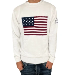 Vit stickad tröja med USA-flagga GANT - Vit stickad tröja från GANT med broderad amerikansk flagga på bröstet och mörkblå text på ena ärmen. Tröjan har rund hals, ribbade muddar och lång ärm. Perfekt för dig som gillar klassisk college-stil och snygga detaljer.