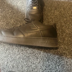 Svarta Nike Air Force 1 sneakers - Säljer ett par svarta Nike Air Force 1 sneakers med klassisk låg siluett. Skorna har snyggt skinnmaterial, perforerad tå och svart snörning med metall-detalj. Perfekt för dig som vill ha en clean och tidlös look med streetkänsla.