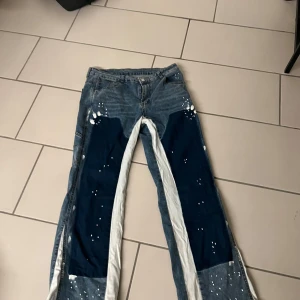 Unika blå jeans med vita detaljer - Säljer ett par riktigt coola jeans med breda ben och kontrasterande mörkblå och vita paneler framtill. Jeansen har vita färgstänk över hela och stora fickor bak samt på sidorna. Materialet är klassisk denim och passformen är loose med baggy vibe.