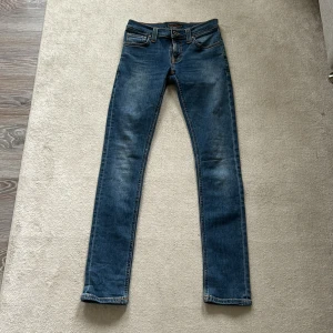 Blå jeans från Nudie Jeans - Snygga blå jeans från Nudie Jeans med klassisk orange söm och broderade bakfickor och storlek 25/32Modellen är skinny med smal passform och normal midja. Jeansen har fem fickor och är tillverkade i mjukt bomullsmaterial med lite stretch för extra komfort.