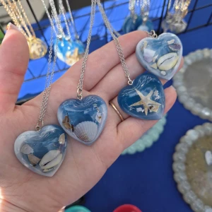 Halsband med hjärta och snäckor - Unika halsband med hjärtformade hängen i blått och vitt, fyllda med små snäckor och en sjöstjärna. Perfekt för dig som älskar strandvibbar och vill ha något som sticker ut. Hängena har en silverfärgad kedja och varje hjärta har sitt eget strandmotiv inuti. Handgjorda med kärlek, skriv vilken du vill ha vid köp.