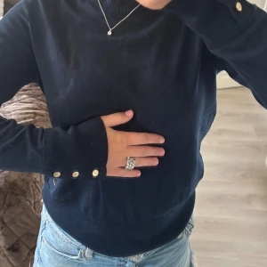 Mörkblå stickad tröja med knappar - Snygg mörkblå stickad tröja från Lindex med rund hals och långa ärmar. Tröjan har guldfärgade knappar vid ärmsluten och en liten metalllogga vid midjan. Perfekt till jeans och smycken för en chill look. Den är knappt använd och kommer inte till användning därav säljer jag den💕