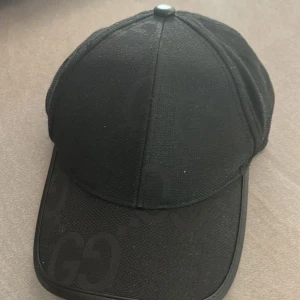 Svart Gucci keps  - Snygg svart keps från Gucci med diskret GG-logga i ton-i-ton mönster över hela kepsen. Både skärm och bakre rem har detaljer i svart skinn. Materialet är bomull och polyester, vilket gör den både bekväm och stilren. Perfekt för dig som gillar exklusiva accessoarer.