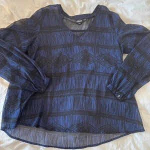 Blå mönstrad blus från Kappahl - Säljer en blå och svart blus från Kappahl med grafiskt mönster och v-ringad hals. Blusen har långa ärmar med knapp vid ärmslut och är gjord i ett lätt, transparent material som ger en luftig känsla. Perfekt för dig som gillar snygga detaljer och avslappnad passform.