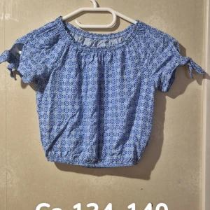 Blå blommig blus med knytärmar - Supersöt blå blus med vitt blommigt mönster och elastisk halsringning. Korta ärmar med knyt i sidan ger en chill och feminin vibe. Perfekt till jeans eller kjol för en fräsch look. Materialet känns lätt och skönt mot huden.