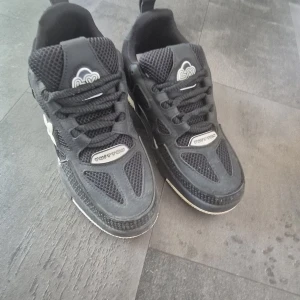 Svarta Louis Vuitton sneakers - Säljer ett par svarta Louis Vuitton sneakers med coola vita detaljer och LV-logga på både sula och häl. Skorna har mesh och skinn i ovandelen, snörning och platt sula. Snyggt mönster på sidan och tydlig branding gör dem till ett statement i din outfit.