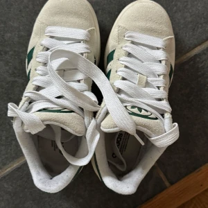 Adidas Campus sneakers i beige och grönt - Säljer ett par Adidas Campus sneakers med beige mocka-överdel och gröna klassiska stripes på sidorna. Skorna har vit snörning, rund tå och en platt gummisula i ljusbrunt. Perfekt för dig som gillar retrostil och vill ha något som sticker ut. 
