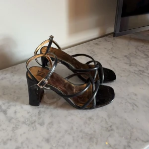 Svarta klackskor - PRIS KAN DISKUTERAS!!! Snygga svarta sandaler från XIT med bred klack och smala remmar som korsar över foten. Med spännband som är justerbart. Klackarna har en fyrkantig tå och justerbart spänne vid ankeln. Insidan av skorna är guldiga. Remarna kan va lite slitet vid spännbandet. Klackarna är cirka 5-6cm. Endast använda en gång på bal. Storlek 36 passar säkert 35 också.