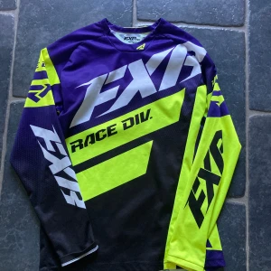 FXR långärmad cykeltröja  - Säljer en riktigt snygg långärmad träningströja från FXR i neon-gult, svart och lila. Tröjan har stora loggor och coola grafiska detaljer, perfekt för dig som gillar att sticka ut. Tillverkad i 100% polyester som andas.