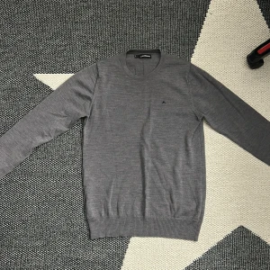 Grå stickad tröja från J.Lindeberg - Snygg grå stickad tröja från J.Lindeberg med rund hals och diskret logga på bröstet. Tröjan har långa ärmar och ribbade muddar vid ärmslut och nederkant. Perfekt för dig som gillar stilrena och klassiska plagg med en modern touch.